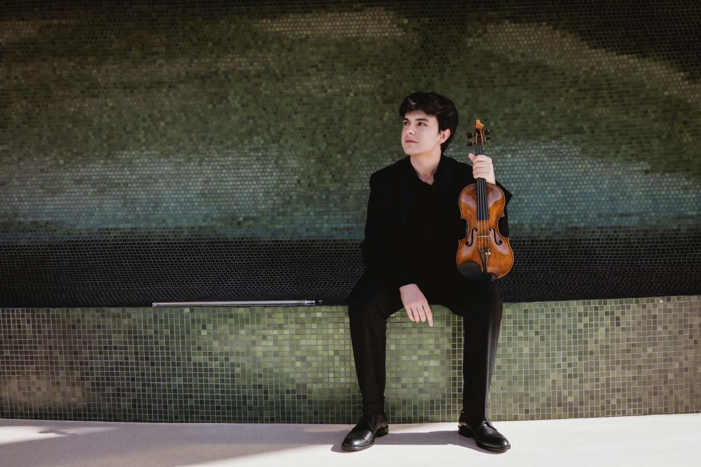 Gabriel Rostagni assis devant une mosaïque verte avec son violon